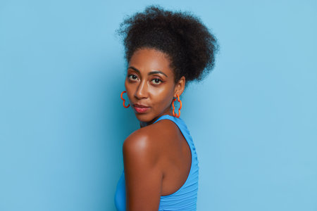 Close-up face to shoulder of beautiful coloured girl in blue top on blue background, serious look into camera, natural beauty concept, copy spaceの写真素材