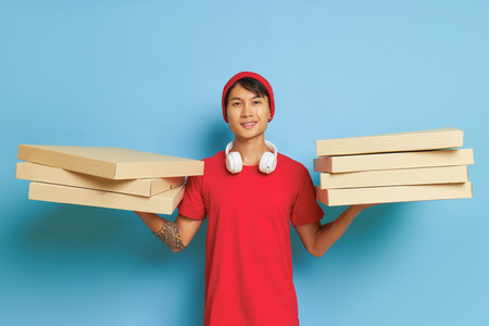 Joyful man in red hat and red t-shirt standing on blue background with pizza boxes in both hands, tasty food concept, copy spaceの写真素材