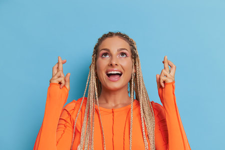 Close-up shot of pretty woman wearing orange longsleeve top on blue background, smiling and crossing her fingers for luck, happy time concept, copy spaceの写真素材