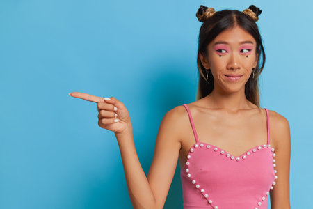 Chinese young woman with stylish makeup in short pink top standing isolated in studio, pointing finger aside, good choice concept, copy spaceの写真素材
