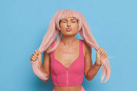Doll style girl in pink top posing on blue background, holding her long pink ponytails in hands and sends airkiss, barby trend concept, copy spaceの写真素材