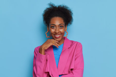 African woman in pink top poses against blue backdrop, holds hand under her chin with smile, lucky time concept, copy spaceの写真素材