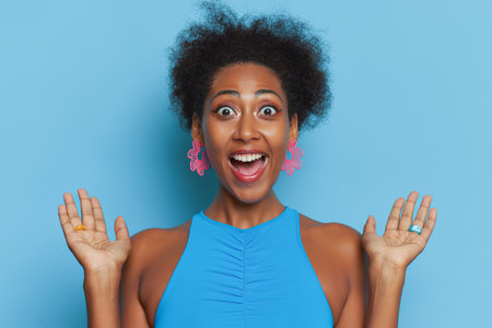 Happy face black woman with curly haircut wearing blue top against blue background with wide smile, good day concept, copy spaceの写真素材