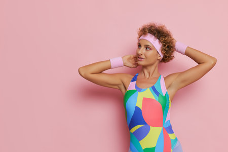 Pretty sportswoman with short curly hair wearing colorful leotard standing on pink background aside with hands over her head, sport lifestyle concept, copy spaceの写真素材
