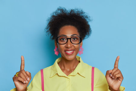 Portrait of smiling coloured girl in glasses against blue background withindex fingers pointed up, decision time concept, copy spaceの写真素材