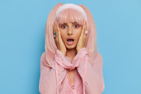 Surprised girl in pink trendy top with long pink hair stands on blue background with hands up on her face, doll trend concept, copy spaceの写真素材