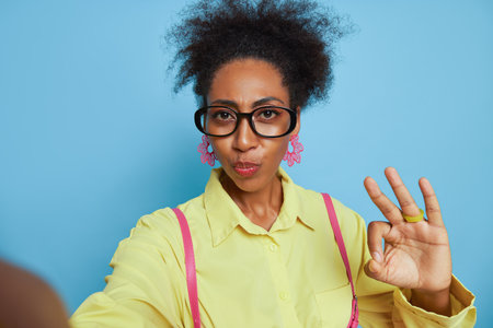 Selfie of black girl in glasses and yellow shirt on blue background showing ok gesture with fingers, interesting facts concept, copy spaceの写真素材