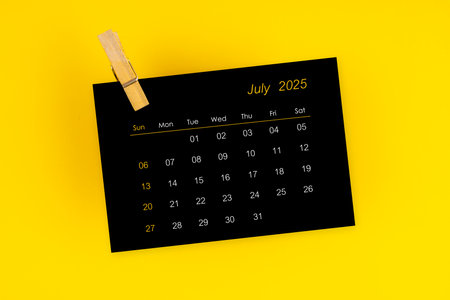 July 2025 black calendar with wooden clip on yellow color background.
