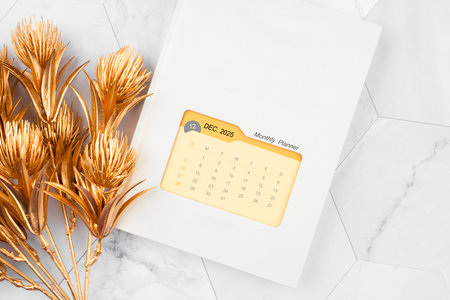 December 2025 calendar on monthly planner book with golden flower on white marble background.