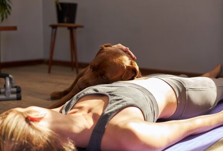 Young female lying on mat after workout at home with dog in the morning sun.