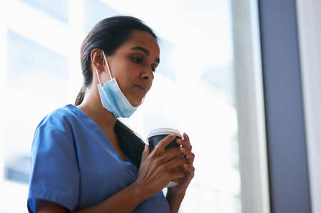 Overworked Nurse In Scrubs With Face Mask Takes Coffee Break In Busy Hospital During Health Pandemicの写真素材