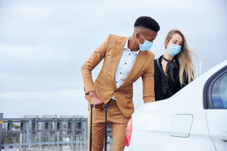 Business Couple Wearing Masks Outside Airport Getting Luggage Out Of Car During Health Pandemicの写真素材