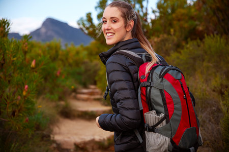 Portrait of young smiling woman on vacation looking over her shoulder wearing backpack walking along...