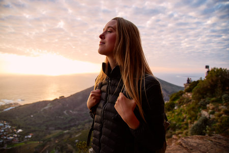 Portrait of smiling young woman on vacation closing her eyes as she walks along coastal path and sun sets over sea behind herの写真素材