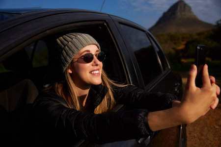 Young woman wearing sunglasses and beanie hat on road trip vacation taking photo out of rental car window on mobile phoneの写真素材