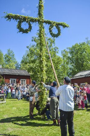 Umea, Sweden - JUNE 23, 2017 Happy people enjoying to watch pole standing moment effort by swedish people and waiting for the swedish mid summer day celebration with colourful environment and blue sky in the background with trees aroundのeditorial素材