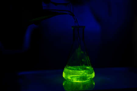 A researcher working with a green fluorescent solution in a glass conical flask in dark biotech...