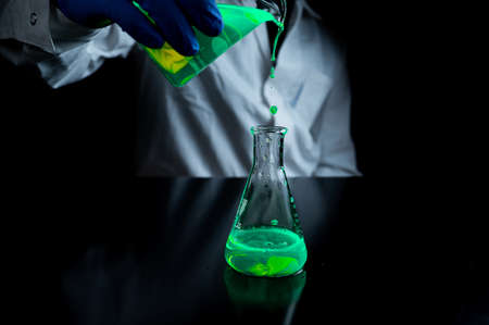 A research scientist experimenting with a green fluorescent droplets in a glass conical flask in dark biomedical laboratory for health care medicine development. Copy space black backgroundの写真素材