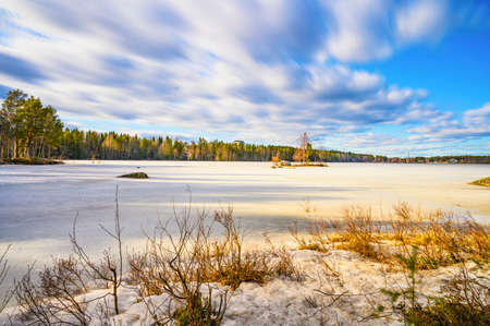 A tranquil landscape of sun lit cloudscape over a frozen lake surrounded by pine tree during the day time in nordic country Swedenの写真素材
