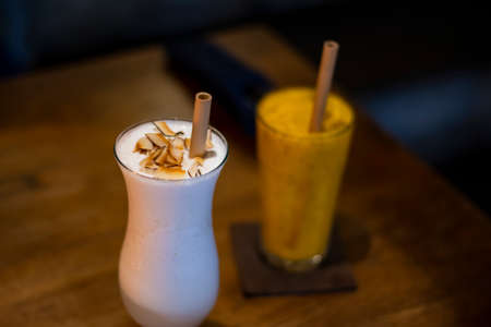 A glass of milk shake with dry coconut on the top and mango smoothie with wooden straw for environment friendly restaurantの写真素材