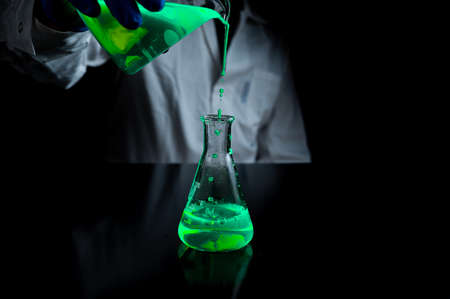 A woman scientist experimenting with a green fluorescent solution in a glass conical flask in dark chemistry laboratory for health care medicine development. Copy space black backgroundの写真素材