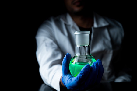 A woman scientist experimenting with a green fluorescent solution in a glass round bottom flask in dark chemistry laboratory for health care medicine development. Copy space black backgroundの写真素材
