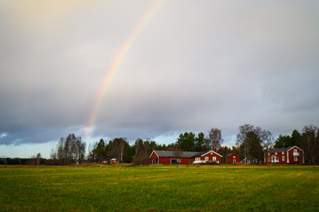 A cloudy landscape view of open field filled with grass with red wooden cottages and rainbow in the sky in a rainy day during a outdoor visit during the springの写真素材