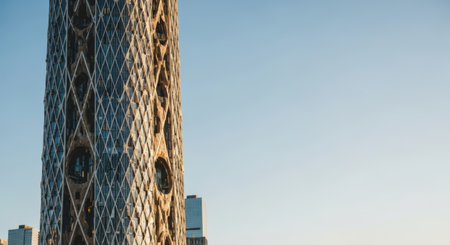 Architectural shot of a modern skyscraper featuring a distinctive diamond-patterned facade with circular openings, set against a clear blue sky. Another building is visible in the background.の素材