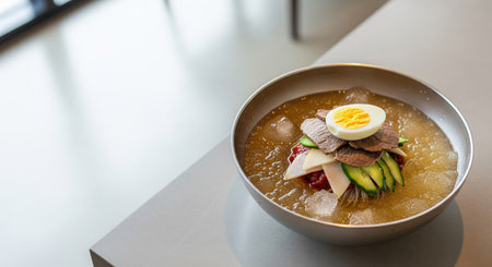 A bowl of Korean Naengmyeon, featuring buckwheat noodles in chilled broth, topped with beef slices, cucumber, pear, egg, and gochujang sauce. Served in a metal bowl with ice.の素材