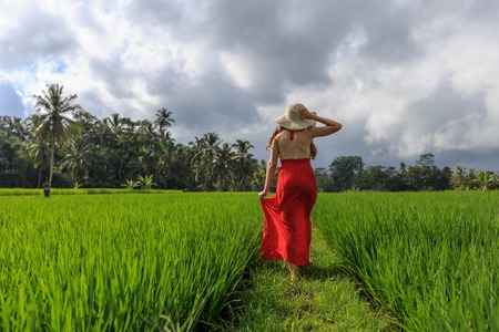 Beautiful young woman in red dress touch straw hat. Girl walk at typical Asian hillside with rice farming, mountain shape green cascade rice field terraces paddies. Ubud, Bali, Indonesia.の写真素材