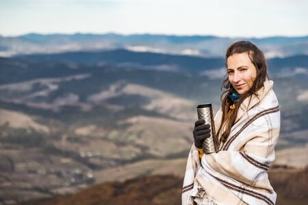 Scenery autumn sunny day. Woman standing covered in plaid with termo cup in hand watching the mountains peak, sky with clouds.の写真素材