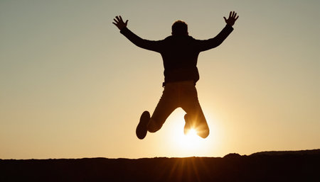 A man's silhouette is seen jumping with arms raised high against a bright sunset. Clear details and vibrant colors en...の素材