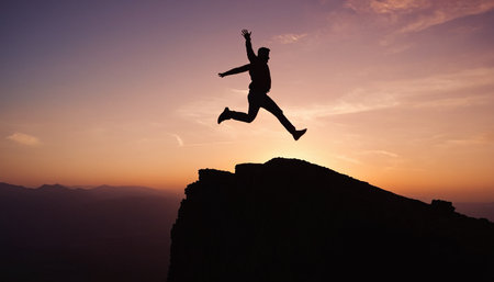 Silhouette of a man leaping from a rocky cliff edge against a vibrant sunset sky. Clear details and vibrant colors en...の素材