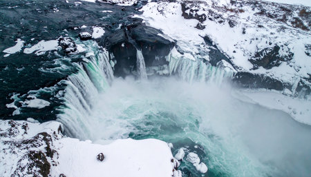 Aerial view of a powerful waterfall in Iceland with snow covered cliffs and turquoise water.の素材