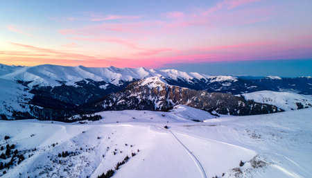 A wide aerial view of a snowy mountain landscape with scattered pine trees and a pink sky.の素材