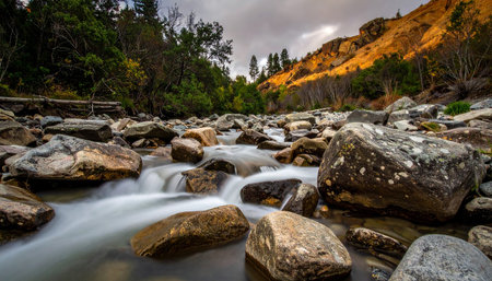 A river flows over rocks with sunlit golden hills and trees in the background. Clear details and vibrant colors enhan...の素材