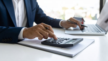 A man in a suit calculates with a calculator and pen near a house model and laptop.の素材