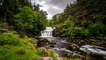 A scenic waterfall flows through a lush green landscape with trees, rocks, and distant hills.の素材