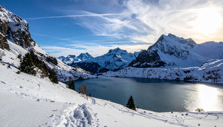 Snowy mountain landscape with footprints leading towards a lake and distant snow covered peaks under a blue sky.の素材