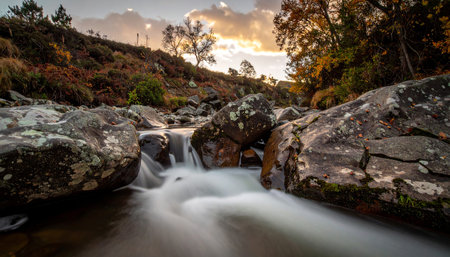 An autumn mountain stream with silky water flowing around large, lichen-covered rocks under a cloudy sky.の素材