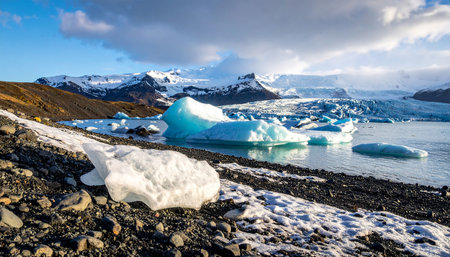 Bright blue icebergs drift in a glacial lagoon with snow-capped mountains under a cloudy sky.の素材