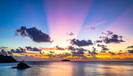 Dramatic crepuscular rays emanate from a colorful sunset sky over a calm ocean dotted with distant tropical islands.の素材