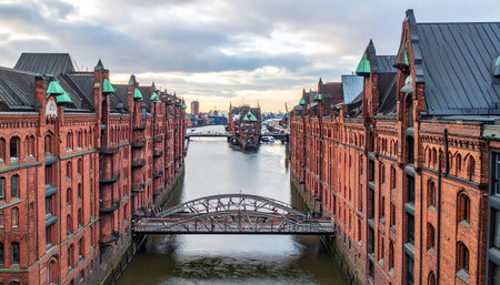 Historic red brick warehouses line a canal in Hamburg Germany with a bridge crossing the water under a cloudy skyの素材