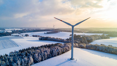 A wind turbine with its blades angled upwards stands on a snow-covered hill with a distant landscape.の素材