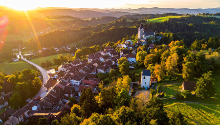 A European village is surrounded by rolling hills and colorful autumn trees during a warm sunset.の素材