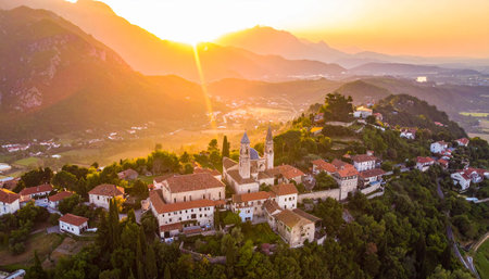Drone view of a European hillside village at sunrise, with church spires and buildings illuminated by warm golden light.の素材
