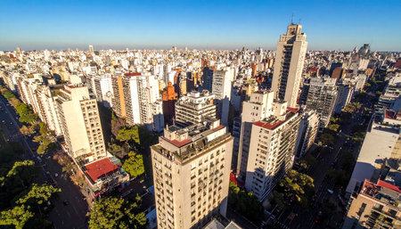 Aerial view of a dense urban cityscape with numerous tall buildings and scattered green trees under a clear blue sky.の素材