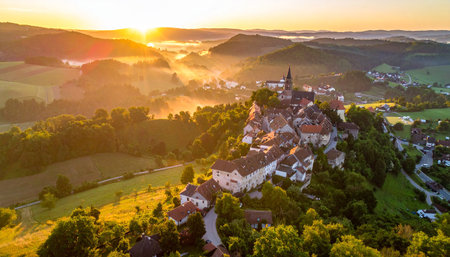 An ancient village sits atop a hill, bathed in the warm glow of sunrise with mist swirling around the landscape.の素材