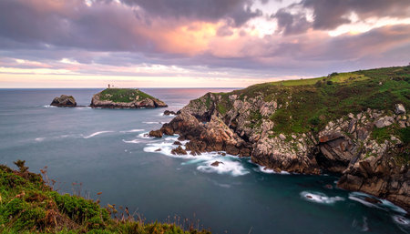 Distant lighthouse on an island with rugged coastline and a moody, cloud-filled sunset sky.の素材