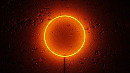 A vibrant yellow neon circle glows intensely against a dark, rough-textured wall. The wall appears to be made of concrete or a similar with an uneven, pitted surface that creates deep shadows and highlights. The neon light casts a warm, orange-red glow onto the wall around the circle, creating a strong contrast between the bright, clean lines of the neon and the ruggedness of the background. The overall effect is modern, and visually striking.の素材